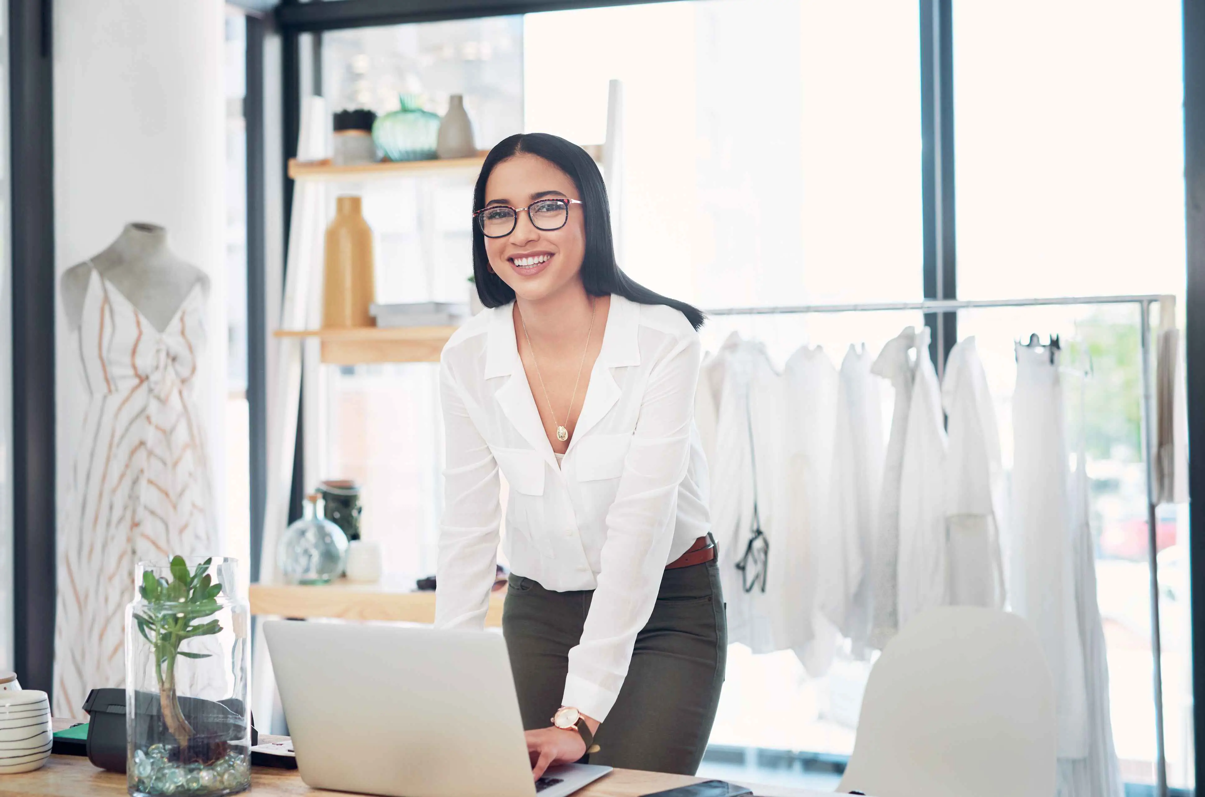 Een dame met een bril en een witte blouse die achter een laptop staat met op de achtergrond witte kledij op een rekje