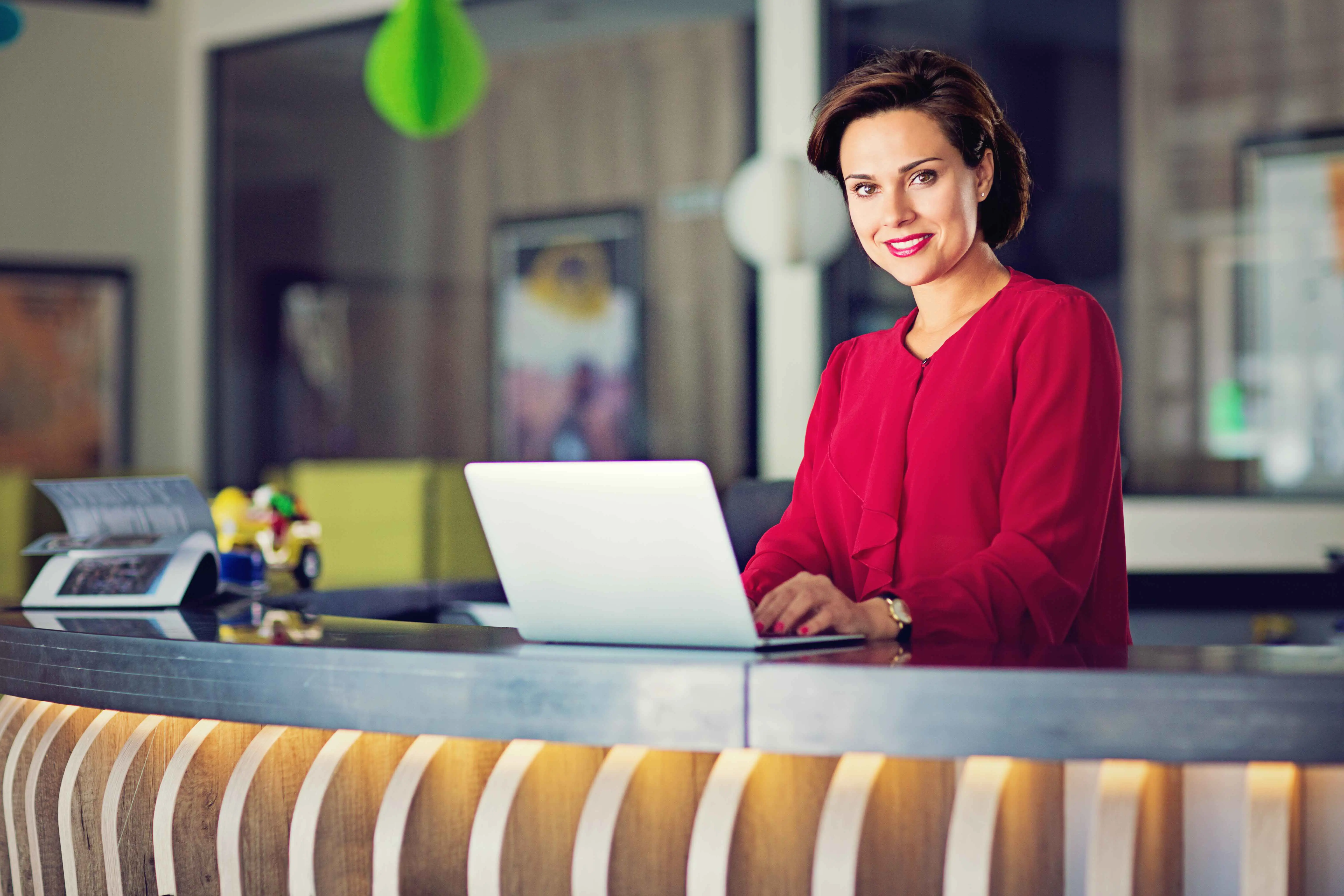 Receptioniste die poseert achter een desk met een laptop. Ze heeft een rode trui  aan 