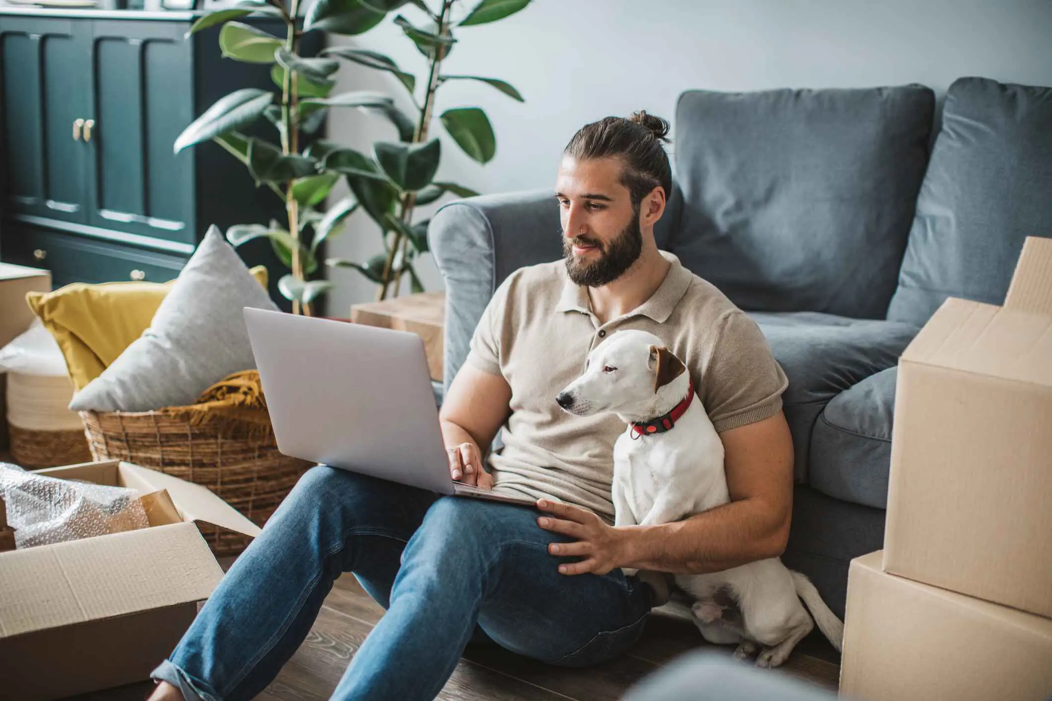 Een man met haar in een staartje zittend op de grond samen met zijn witte hond kijkend naar een laptop